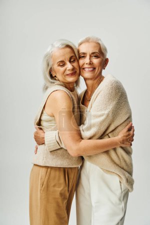 senior lifelong female friends in fashionable casual attire embracing on grey, sophisticated aging