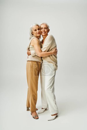 Photo for Full length of elegant and happy mature women in stylish pastel clothes hugging each other on grey - Royalty Free Image