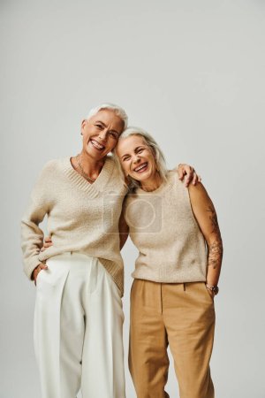 Photo for Cheerful mature ladies in trendy pastel embracing and looking at camera on grey, lifelong friends - Royalty Free Image