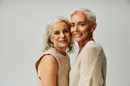 modern senior female friends in pastel clothes smiling at camera on grey, happy and stylish aging