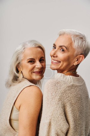 mujeres mayores alegres y elegantes con el pelo plateado sonriendo en gris, felicidad y envejecimiento de moda
