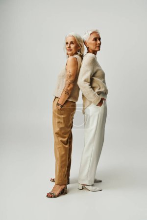volle Länge selbstbewusster Seniorinnen, die Rücken an Rücken mit den Händen in den Taschen vor grauem Hintergrund stehen