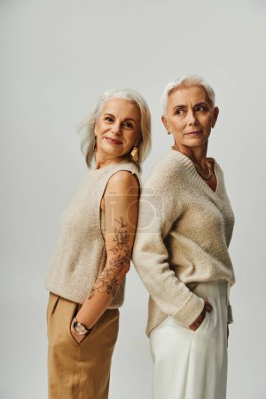 Senior tätowierte Frau lächelt in die Kamera neben stilvolle Freundin auf grau, altert anmutig