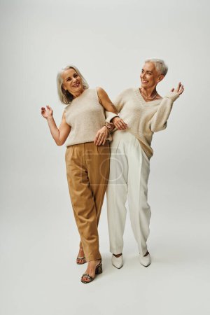 volle Länge der aufgeregten und modischen Seniorinnen, die Spaß auf grauem Hintergrund haben, positives Altern
