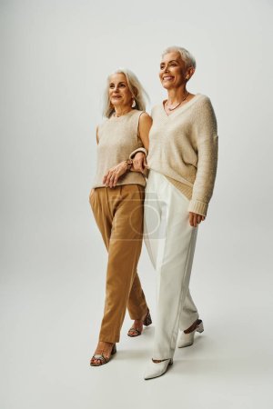 fashionable and happy senior female friends walking arm in arm on grey background, full length