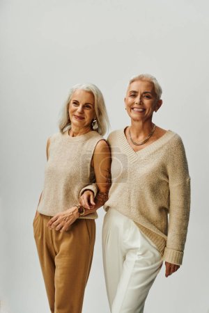 pleased mature women in trendy casual clothes standing arm in arm on grey, fashion and friendship