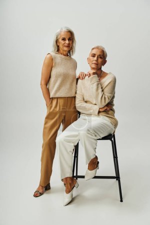 elegant senior woman sitting on chair and looking at camera near smiling female friend on grey