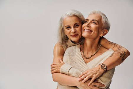 Foto de Alegre mujer tatuada abrazando feliz y de moda amiga en gris, la amistad de los modelos senior - Imagen libre de derechos