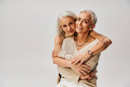 fashionable tattooed woman hugging mature delighted female friend on grey, positive lifestyle