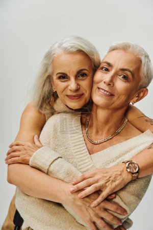 Foto de Alegre y de moda mujer tatuada abrazando complacida madura amiga en gris, amistad atemporal - Imagen libre de derechos