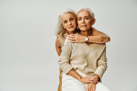 femme tatouée glamour embrassant souriant et élégant ami féminin assis sur gris, personnes âgées à la mode