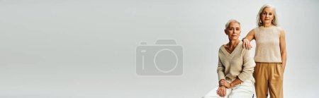 Photo for Mature lady sitting near female friend standing with hand in pocket on grey, senior fashion, banner - Royalty Free Image