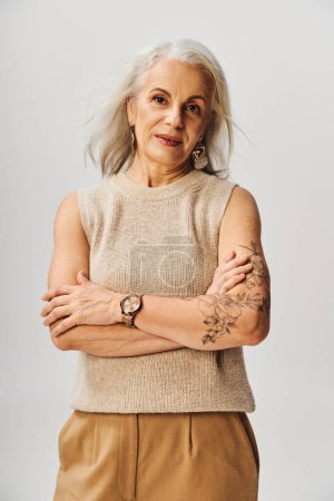 modische und tätowierte Seniorin posiert mit verschränkten Armen und blickt in die Kamera auf grau