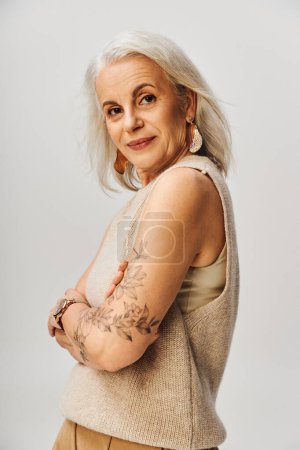 Foto de Señora mayor tatuada y de moda en el oeste de punto posando con los brazos cruzados, sonriendo a la cámara en gris - Imagen libre de derechos