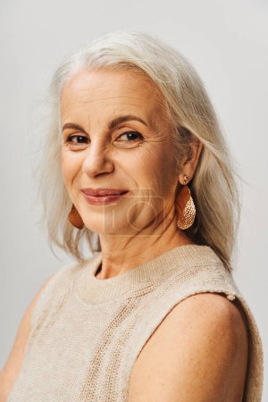 Porträt einer glücklichen Seniorin in Make-up und goldenen Ohrringen, die vor grauem Hintergrund in die Kamera blickt