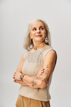 Téléchargez les photos : Dame tatouée mature en haut tricoté debout avec les bras croisés et regardant loin sur gris, positivité - en image libre de droit