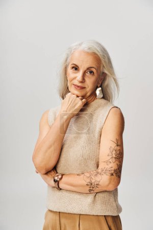 senior tattooed woman in pastel top holding hand near chin and looking at camera on grey backdrop