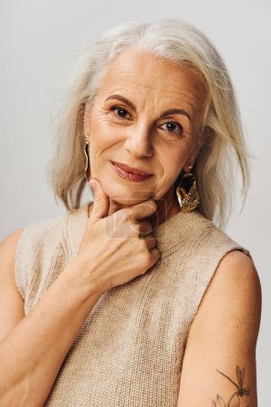 portrait de mannequin senior aux cheveux argentés en maquillage et boucles d'oreilles dorées souriant à la caméra sur gris