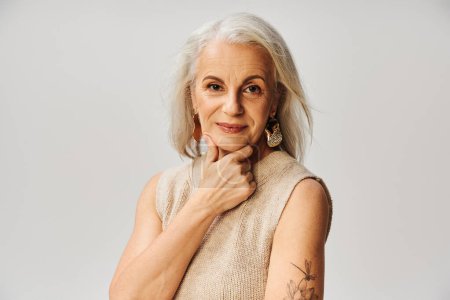 Photo for Portrait of joyful mature lady in makeup, golden earrings and knitted top touching chin on grey - Royalty Free Image