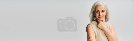 Téléchargez les photos : Élégante femme âgée avec des cheveux argentés et la main près du menton en regardant la caméra sur fond gris, bannière - en image libre de droit