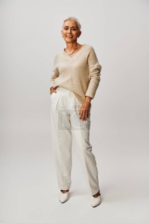 trendige reife Dame im geknoteten Pullover und weißer Hose posiert mit der Hand in der Tasche auf grau, in voller Länge