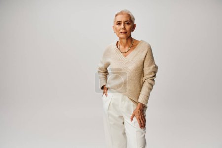 confident senior fashionista with silver hair posing with hand in pocket of white pants on grey