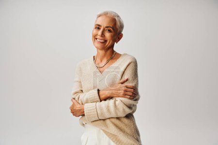 joyful senior model in soft knitted jumper posing with folded arms and smiling at camera on grey