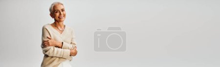 fashionable and smiley mature lady with folded arms looking at camera on grey backdrop, banner