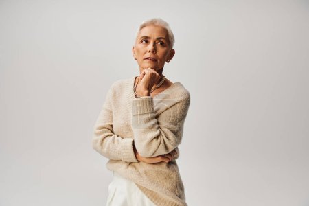 femme âgée rêveuse en tenue décontractée tendance tenant la main près du menton et regardant loin sur gris
