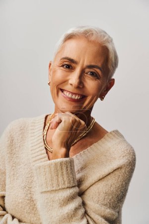 portrait of smiling senior lady with natural makeup and golden accessories looking at camera on grey