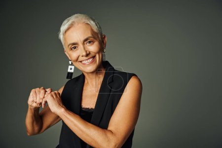 glückliche ältere Dame in stylischen Ohrringen und schwarzer Kleidung lächelt in die Kamera auf grau, Eleganz und Charme