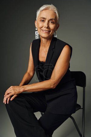 joyful and charming mature lady in black elegant attire sitting on chair and smiling on grey