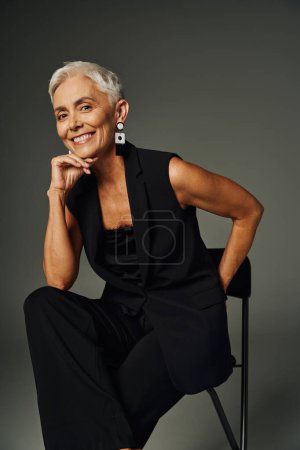 positive, cheerful senior woman in black attire sitting on chair with hand behind back on grey
