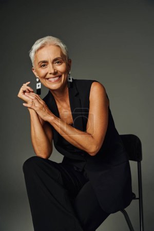charming and happy senior lady in black classic attire looking at camera and posing on chair on grey