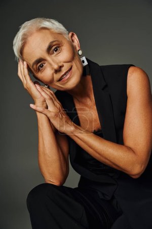 glückliche Seniorin mit kurzem silbernem Haar sitzt in schwarzer Kleidung und blickt in die Kamera auf grau, Portrait