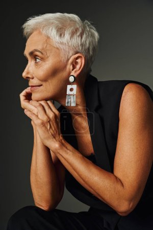 Photo for Portrait of dreamy and positive senior lady with short silver sitting in and looking away on grey - Royalty Free Image