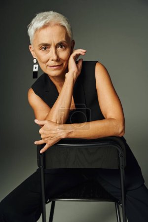 Charmante Seniorin in schwarzer klassischer Kleidung sitzt auf einem Stuhl und blickt in die Kamera auf grau