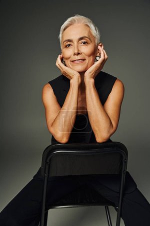 anmutige Seniorin in schwarzer klassischer Kleidung posiert auf Stuhl mit Händen in Gesichtsnähe auf grau
