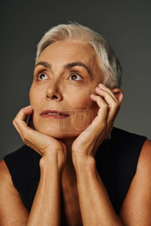 Porträt einer verträumten Seniorin mit kurzen silbernen Haaren, die mit den Händen in Gesichtsnähe auf grau wegschaut