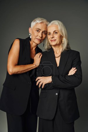 Foto de Vanidad estilo justo, mujeres mayores elegantes en ropa elegante negro de pie sobre fondo gris - Imagen libre de derechos