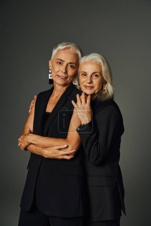 Téléchargez les photos : Positif mature dames en noir élégant vêtements regardant caméra sur gris, vieillissement gracieusement - en image libre de droit