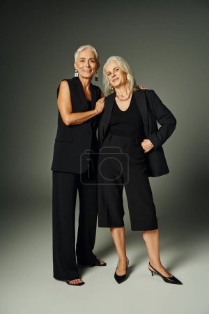 joyful senior female friends in black stylish attire looking at camera on grey, fashionable aging