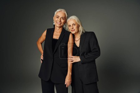pleased senior models in black fashionable clothes looking at camera on grey, charm and elegance