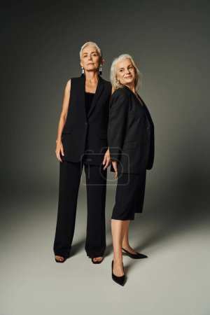 full length of graceful elderly women in black stylish clothing standing on grey, classic fashion