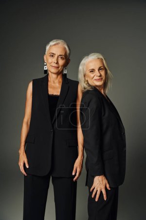 positive mature models in black classic attire smiling at camera on grey, timeless elegance