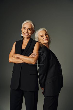 Foto de Alegre mujer madura con los brazos cruzados cerca de sonriente amiga en gris, seniors de moda - Imagen libre de derechos