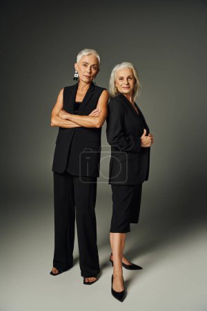 Foto de Moda señora mayor en traje negro posando con los brazos cruzados cerca de amiga femenina en gris, elegancia - Imagen libre de derechos