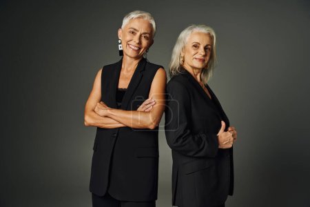 delighted senior woman in black clothes posing with folded arms near female friend on grey backdrop