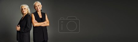 happy mature lady in black classic attire posing with folded arms near female friend on grey, banner