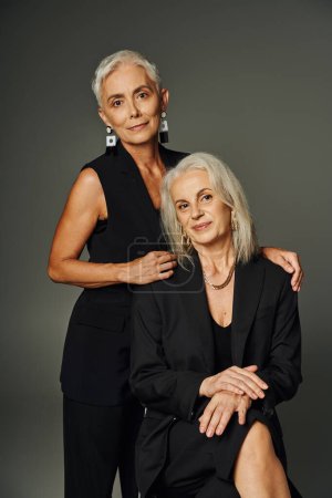 fashionable silver haired woman hugging shoulders of elegant female friend sitting on grey